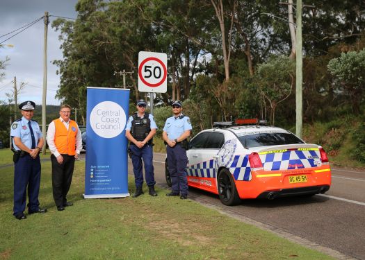 Central Coast Council Director stands alongside three NSW Police Officers in uniform, a 50 kilometre per hour sign and a NSW Police car on the roadside