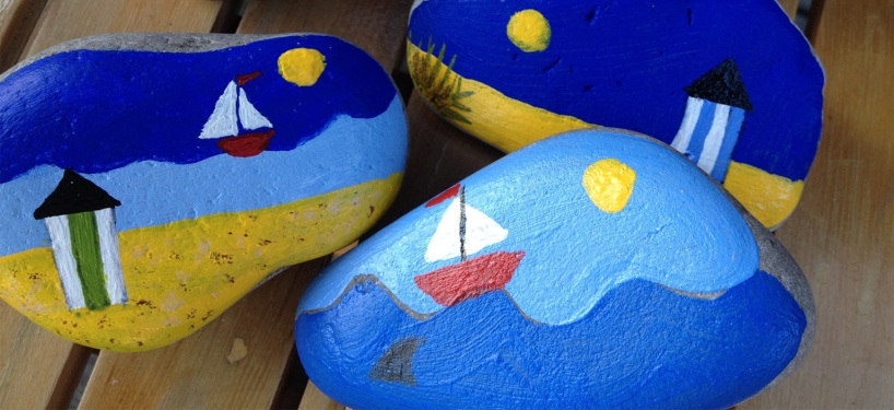 Three small stones, hand decorated with painted images of seaside themes including water, beach, boats and sun