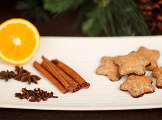 White rectangular plate holding cinnamon sticks, star anise, half an orange and star shaped biscuits - pine tree cone in background