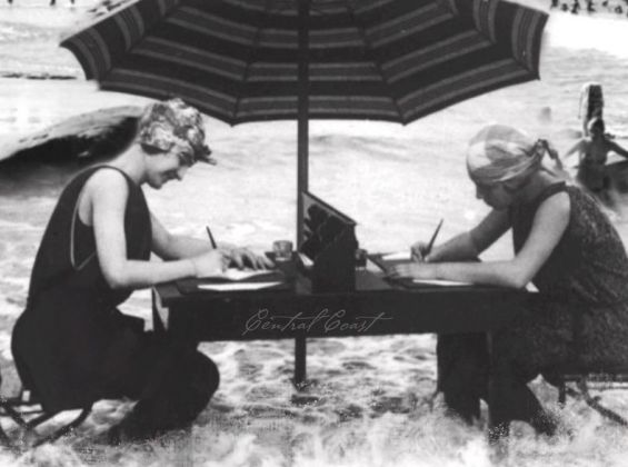 Black and white photo two ladies in the surf sitting at a desk with beach umbrella, writing.
